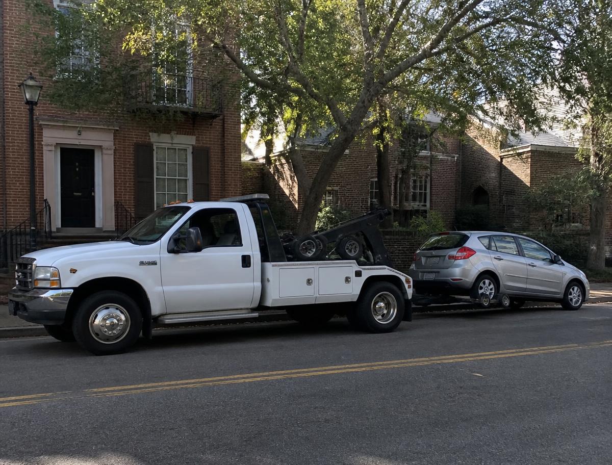 Wheel-lift tow truck towing a hatchback down a tree-lined city street